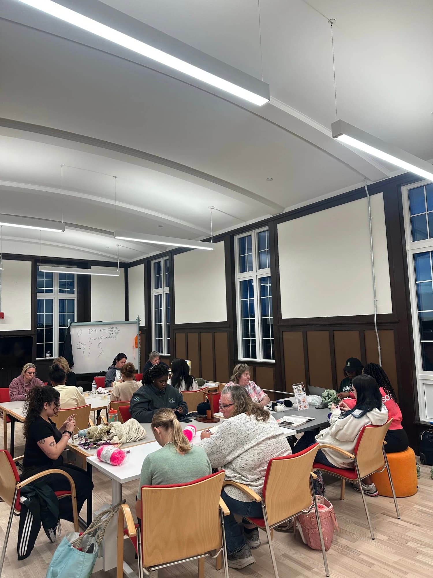 A full room of people crocheting and knitting around long tables at a club meeting